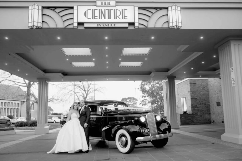 Bride and groom posing next to a vintage car under the illuminated Art Deco entrance porte-cochère at The Centre Ivanhoe.