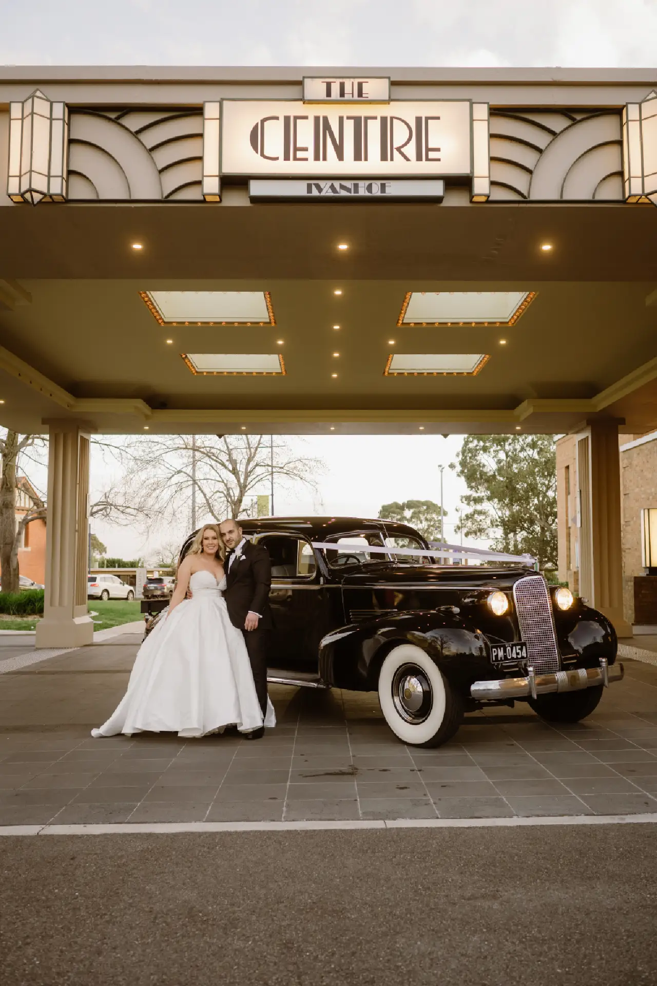 Wedding couple standing next to a black vintage automobile beneath the entrance canopy of The Centre Ivanhoe.